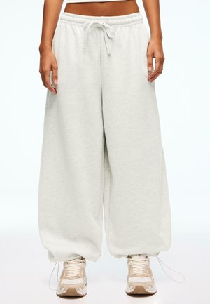 Luźne jasnoszare spodnie dresowe z troczkiem w pasie i elastycznymi ściągaczami, połączone z beżowo-białymi trampkami, noszone przez stojącą osobę.