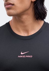 Gros plan d'une personne portant un t-shirt Nike Pro noir avec un logo Nike rose centré sur la poitrine.