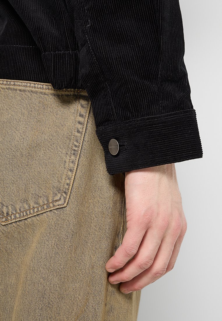 Poignet de veste en velours côtelé noir avec des côtes texturées, arborant un bouton en métal. Pantalon en denim beige visible sur le côté.