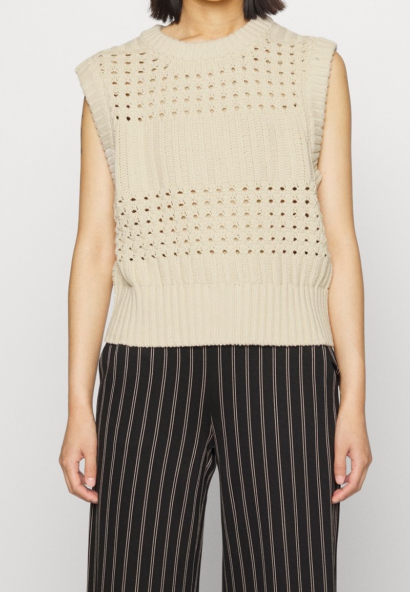 Beige pletený top bez rukávů se strukturovaným vzorem a otevřenými detaily, zakončený žebrovaným lemem, v kombinaci s černými kalhotami s tenkým pruhováním.
