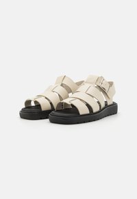 Beige sandalen met bandjes en een zwarte rubberen zool, met een open teenontwerp en een gespsluiting op de enkelband.