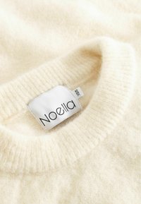 Kremo spalvos megztinis, pagamintas iš minkštos, tekstūruotos medžiagos, su raukinėta iškirpte. Yra prisegtas prekės ženklo "Noella" etiketė.
