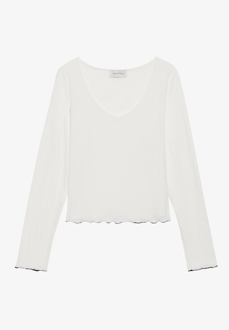 Haut à manches longues blanc avec un décolleté arrondi, texture côtelée et ourlet à volants. Étiquette indiquant "ANNA FIELD."