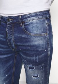 Niebieskie dżinsy o slim fit, z przetarciami na kolanach i białymi plamami farby, z kontrastującymi pomarańczowymi szwami oraz zapięciem na guzik z przodu.