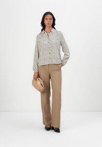 Hvit bluse med svarte prikker, lange ermer og knapper foran. Kombinert med beige bukser og en myk, beige clutch-veske. Svarte flate sko.