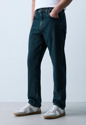 Person trägt dunkelblaue Straight-Leg-Jeans, eine Hand in der Tasche, und weiße Sneaker mit hellgrauen Akzenten.