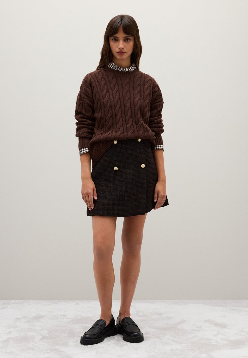 Brauner Kabelstrickpullover über einem schwarzen, strukturierten Minirock mit goldenen Knöpfen, kombiniert mit schwarzen Loafers und einem karierten Kragen.