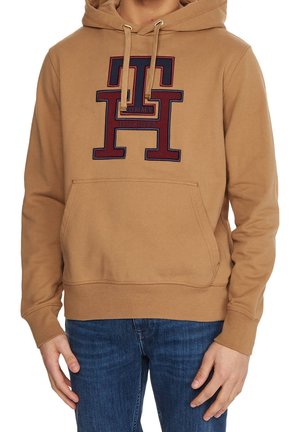 Mężczyzna noszący beżową bluzę z kapturem z dużym wyszywanym logo "TH" i napisem "Tommy Hilfiger" z przodu, w połączeniu z niebieskimi dżinsami.