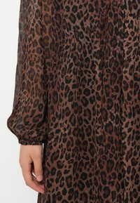 Tuvumā redzama roka, kas balstās uz brūna un melna leoparda raksta plisētām kleitām ar garām, aprocēm caurspīdīgām piedurknēm.