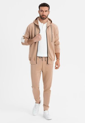Mann läuft, trägt beige Zip-up-Hoodie und passende Jogginghosen, weißes Strickshirt und weiße Sneakers vor einem einfarbigen weißen Hintergrund.