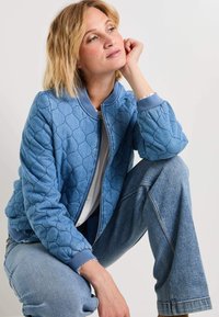 Chaqueta bomber acolchada azul con cuello y puños de canalé, que presenta un cierre de cremallera y un patrón texturizado. Llevada sobre una blusa ligera y denim.