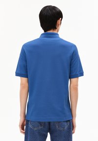 Blaues Polohemd mit kurzen Ärmeln, ausgestattet mit einem klassischen Kragen und strukturiertem Stoff. Getragen mit Jeanshosen, die ein einfaches, lässiges Design zeigen.