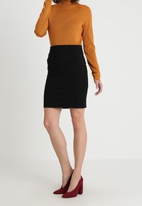 Oranžový třpytivý rolák v kombinaci s černou úzkou sukní. Modelka má na sobě vínové strukturované lodičky, které se vyznačují hladkým, minimalistickým designem.