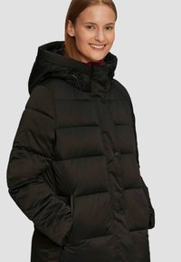 Schwarze Pufferjacke mit Kapuze, ausgestattet mit gesteppter Oberfläche, vorderen Knöpfen und Taschen. Hergestellt aus glattem, wasserabweisendem Material.