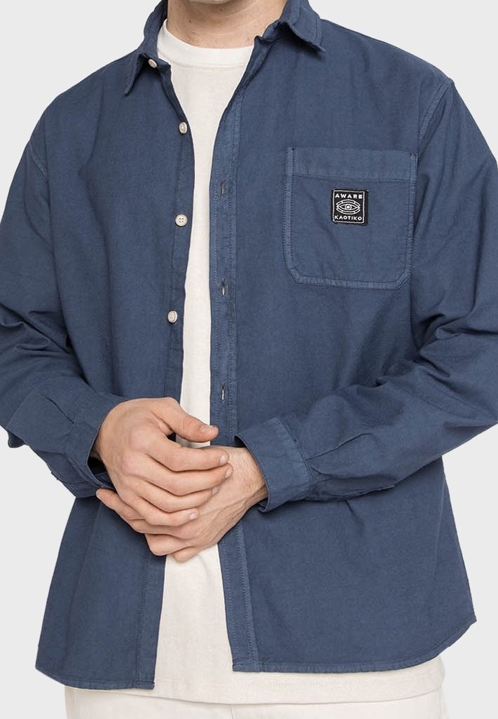Personne portant une chemise bleue déboutonnée avec un patch sur la poche indiquant « Aware Kaotiko » par-dessus un t-shirt blanc, mains jointes à la taille.