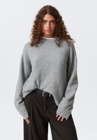 Un pull en maille grise avec une coupe décontractée, des épaules basses et des poignets côtelés, associé à un pantalon sombre et une ceinture noire. Design minimaliste.