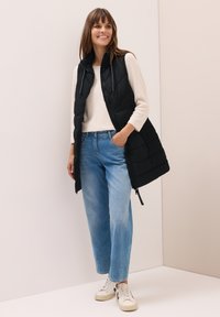 Schwarze gepolsterte Weste mit hohem Kragen über einem beigefarbenen Pullover, kombiniert mit hellblauen, lässig geschnittenen Jeans und weißen Sneakers mit schwarzen Akzenten.
