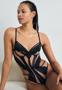 Mustmustel lace bodysuit läbipaistvate paneelidega, millel on sügav kaelus, õhukesed reguleeritavad rihmad ja dekoratiivsed pitsaktsendid. Sile tekstuur.