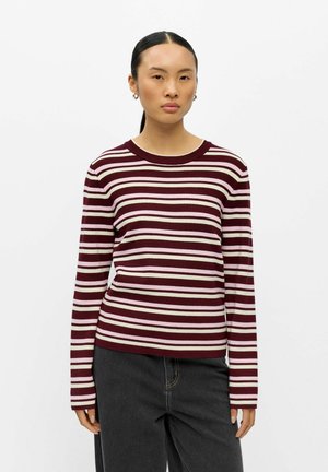 Jeune femme aux cheveux foncés attachés porte un pull rayé à manches longues bordeaux, rose et blanc et un pantalon noir sur un fond uni.
