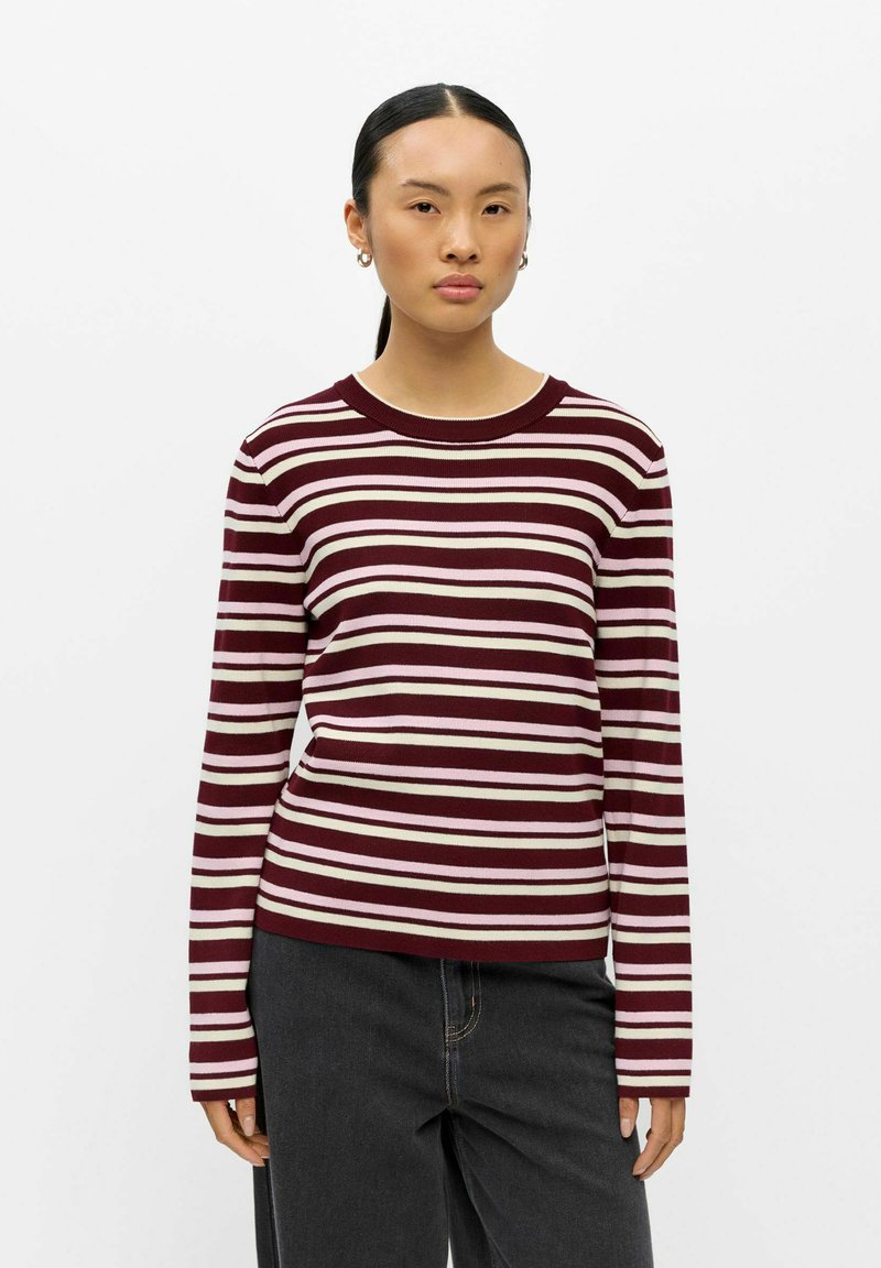 Jeune femme aux cheveux foncés attachés porte un pull rayé à manches longues bordeaux, rose et blanc et un pantalon noir sur un fond uni.