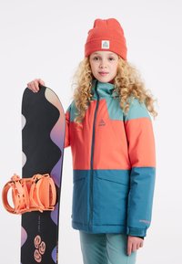 Dijete drži crni snowboard s narančastim vezicama, noseći jaknu u boji koja se sastoji od tirkizne i koraljne, uz odgovarajuću koraljnu kapu.