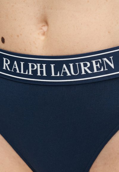 Κοντινή λήψη του κάτω μέρους της κοιλιάς ενός ατόμου που φοράει εσώρουχο μπλε ναυτικού με λευκή ζώνη μέσης με το λογότυπο "Ralph Lauren" και εμφανές ομφαλό.