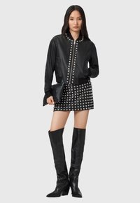 Giacca bomber in pelle nera con borchie argento, abbinata a una mini gonna nera borchiata e stivali neri alti sopra il ginocchio, mentre tiene una pochette nera.