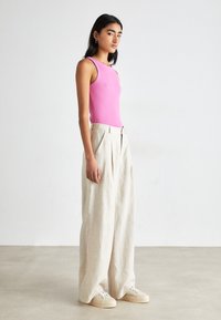 Anpassungsfähiges pinkes Tanktop, hochtaillierte, locker sitzende cremefarbene Leinenhose mit Falten und hellen Sneakers. Schlichter Hintergrund.