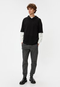 Zwarte hoodie met korte mouwen en een voorzak, aangetrokken over een witte shirt met lange mouwen; grijze cargobroek met verstelbare boorden en zakken.