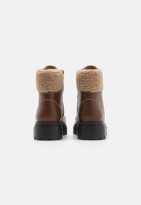 Bottes de cheville en cuir marron avec un col beige moelleux, des semelles en caoutchouc noir épaisses, et une finition lisse sur le corps, avec des détails de couture contrastants.