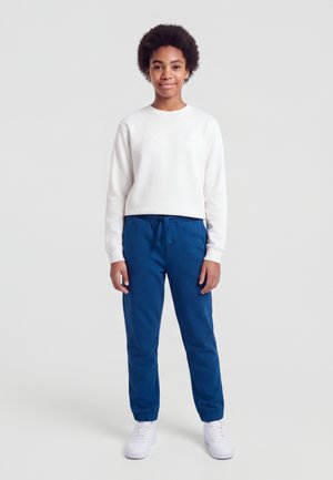 Jeune personne aux cheveux courts bouclés portant un sweat-shirt blanc, un pantalon de jogging bleu et des baskets blanches, debout devant un fond uni.
