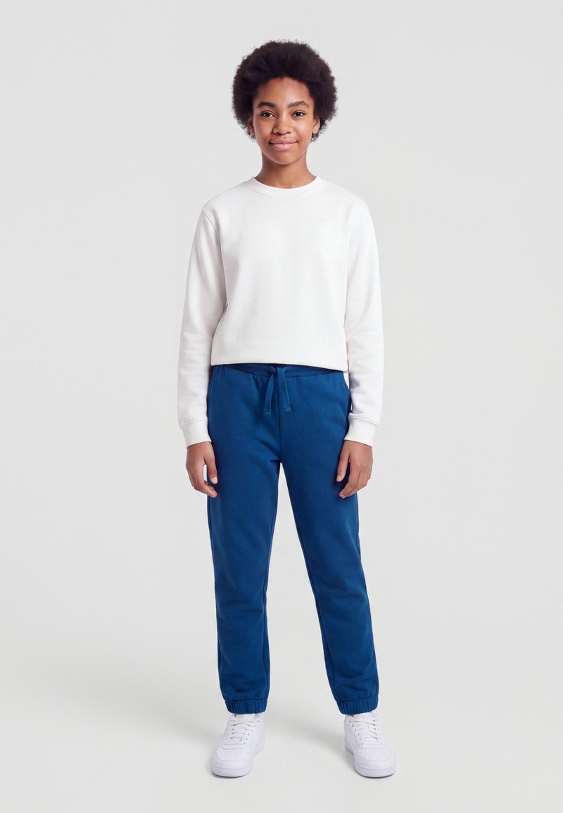 Jeune personne aux cheveux courts bouclés portant un sweat-shirt blanc, un pantalon de jogging bleu et des baskets blanches, debout devant un fond uni.