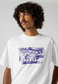 Valkoinen puuvillapaita, jossa on violetti graafinen kuvasto, jossa on ikonisia pariisilaisia kylttejä, mukaan lukien "Metropolitain" ja "Boulangerie" -yksityiskohtia.