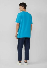 Turquoise short-sleeve t-shirt with a crew neck, paired with dark blue pants and white sneakers, viewed from the back. Smooth texture.