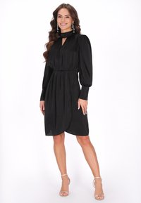 Abito nero con maniche a sbuffo lunghe, scollatura a chiave e vita elasticizzata. Tessuto morbido, orlo a ginocchio, abbinato a sandali con tacco trasparente.