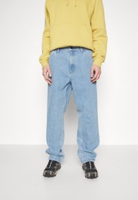 Vaqueros de mezclilla azul claro con un corte relajado, cintura alta y piernas rectas. Combinados con una sudadera con capucha amarilla y zapatos negros.