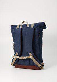 Marineblå rulletop ryggsekk med polstrede stropper, brun bunn, justerbar brystspenne og sidelommer med glidelås. Holdbart stoff og tekstur.