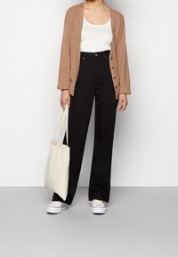 Frau trägt einen braunen Cardigan, ein weißes Tanktop, schwarze High-Waist-Hosen und weiße Turnschuhe. Sie hält eine beigefarbene Tragetasche und steht auf einem grauen Boden.