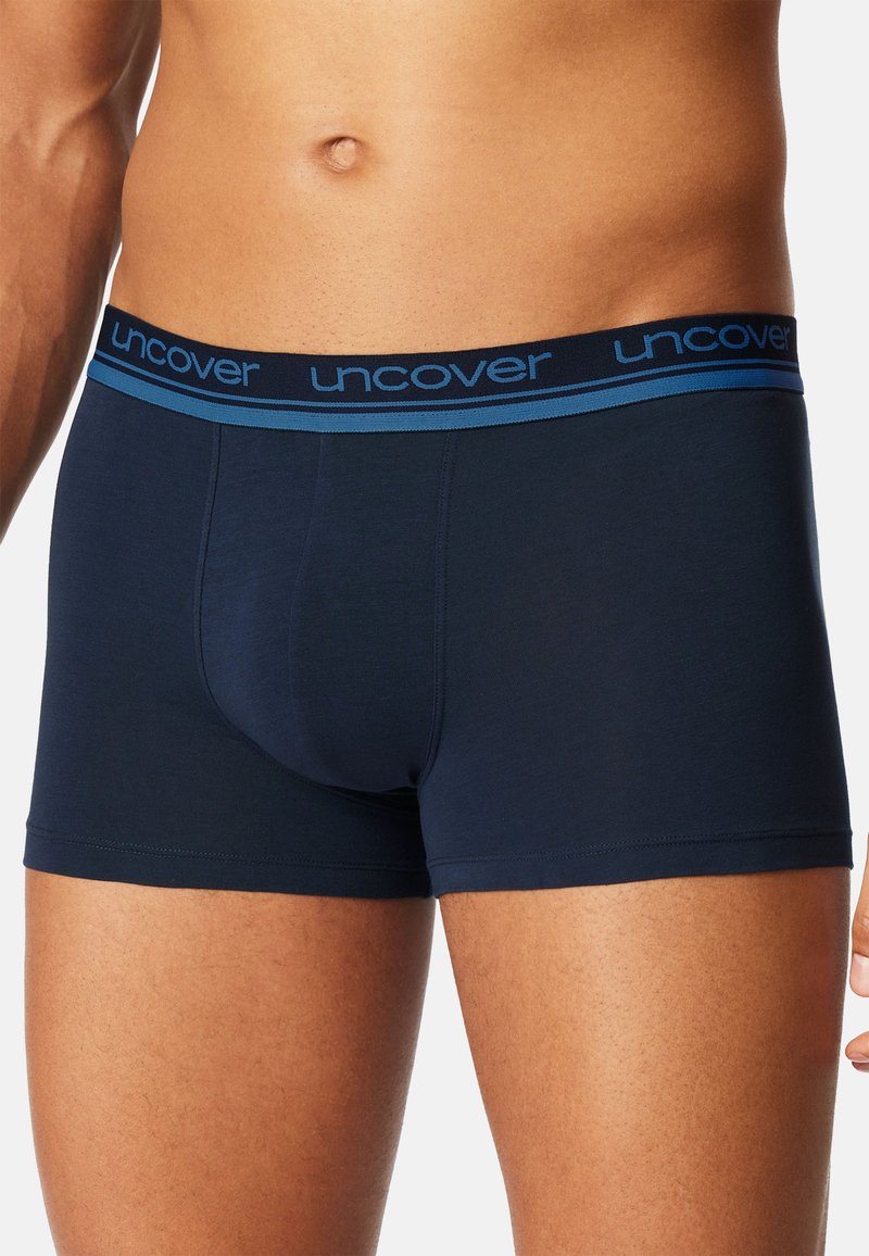 Mannelijke torso gekleed in marineblauwe boxershorts met een blauwe elastische tailleband waarop "uncover" staat, tegen een witte achtergrond.