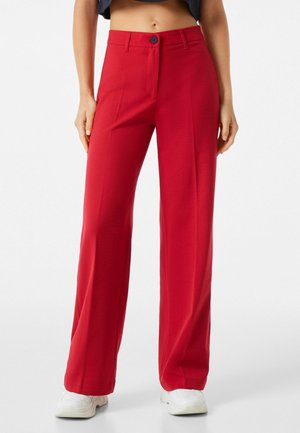 Vrouw die rode broek met hoge taille en wijde pijpen draagt met knoopsluiting, gecombineerd met witte sneakers, staand tegen een witte achtergrond.