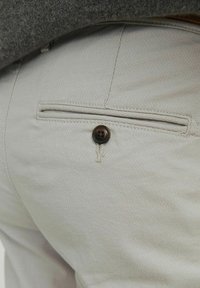 Helle graue Hose aus strukturiertem Stoff, mit einer Gesäßtasche und einem einzelnen braunen Knopf sowie Steppnähten.