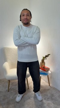 Biely dlhý sveter s textúrou, tmavé rifle a biele tenisky, ktoré nosí osoba stojaca s prekríženými rukami. Svetlé pozadie a nábytok.