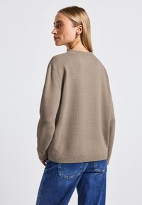 Kvinde med blonde hår iført en taupe strikket sweater og blå jeans, stående med ryggen til og kiggende til siden.
