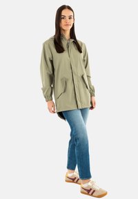 Chaqueta impermeable de color verde claro con capucha, puños elásticos, dos bolsillos laterales, combinada con jeans azules y zapatillas beige con acentos rojos.