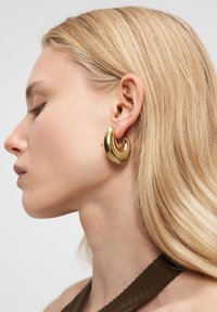 Frau mit langen blonden Haaren, die einen großen, glänzenden goldenen halbmondförmigen Ohrring und ein dunkelbraunes ärmelloses Oberteil trägt, von links angesehen.