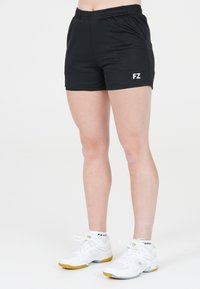 Ben som har svarta träningsshorts med "FZ"-logotyp och vita sportskor med gula sulor, som står på en vit bakgrund.