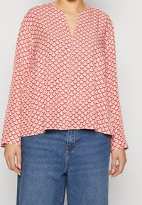 Red and white blouse featuring a scallop pattern, V-neckline, long sleeves, and a loose fit, paired with blue jeans.