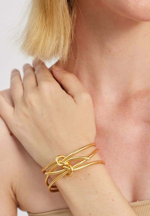 Guldfarvet metalarmbånd med sammenflettede ringe, cylindrisk form, glat tekstur og et enkelt design på bærerens håndled.