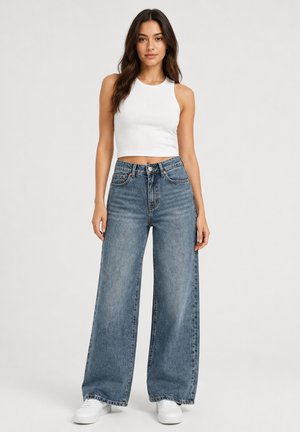 Donna con lunghi capelli scuri che indossa un crop top bianco senza maniche, jeans blu a vita alta e gamba larga e sneakers bianche, in piedi davanti a uno sfondo semplice.