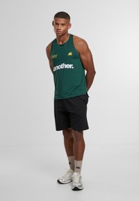 Muž na sobě má zelenou bezrukávovou košili s nápisem "another", černé šortky, béžové ponožky a stříbrné tenisky, stojící s rukama za zády.